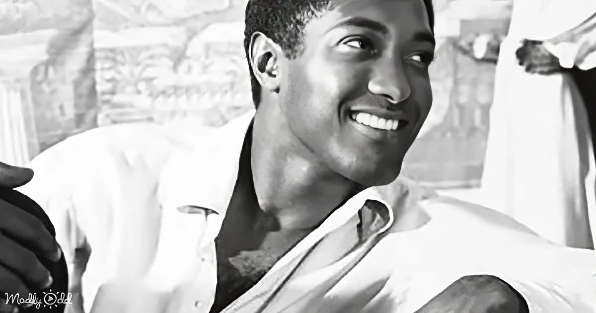 A smiling man in a white shirt, partially unbuttoned, with a relaxed posture and a blurred background.