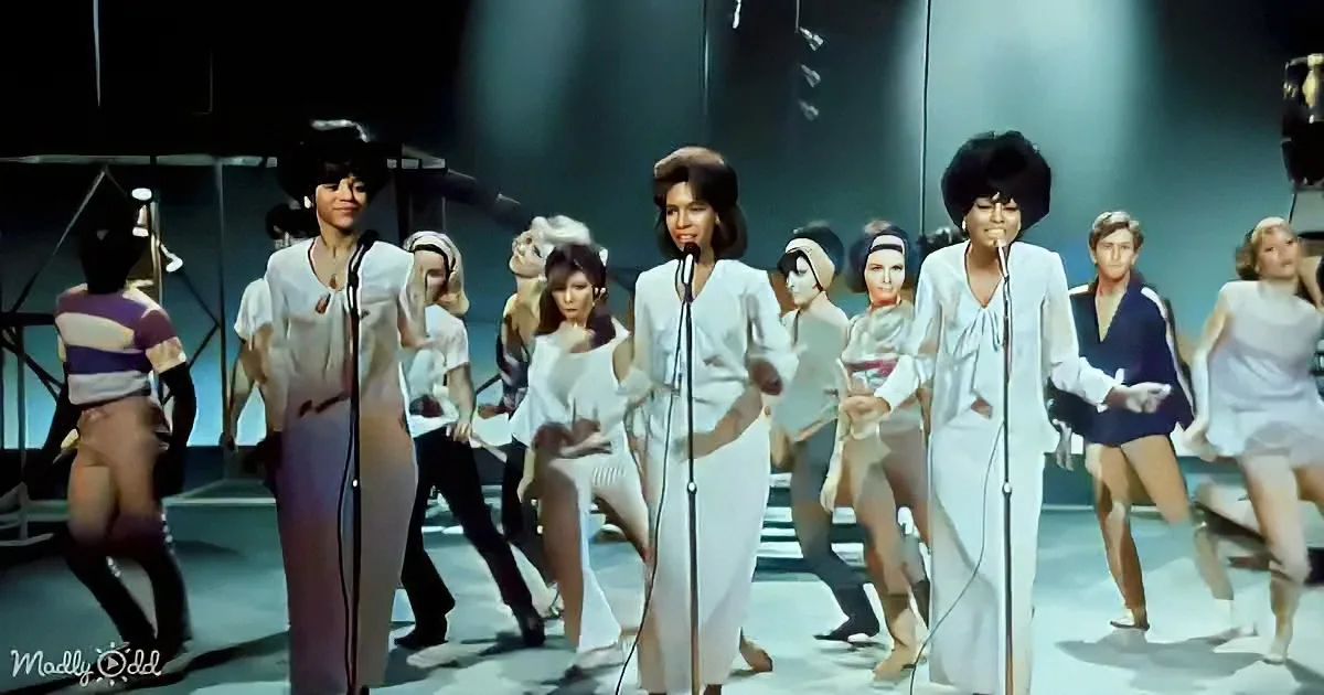 Two women in white outfits sing on stage with a group of dancers in the background.