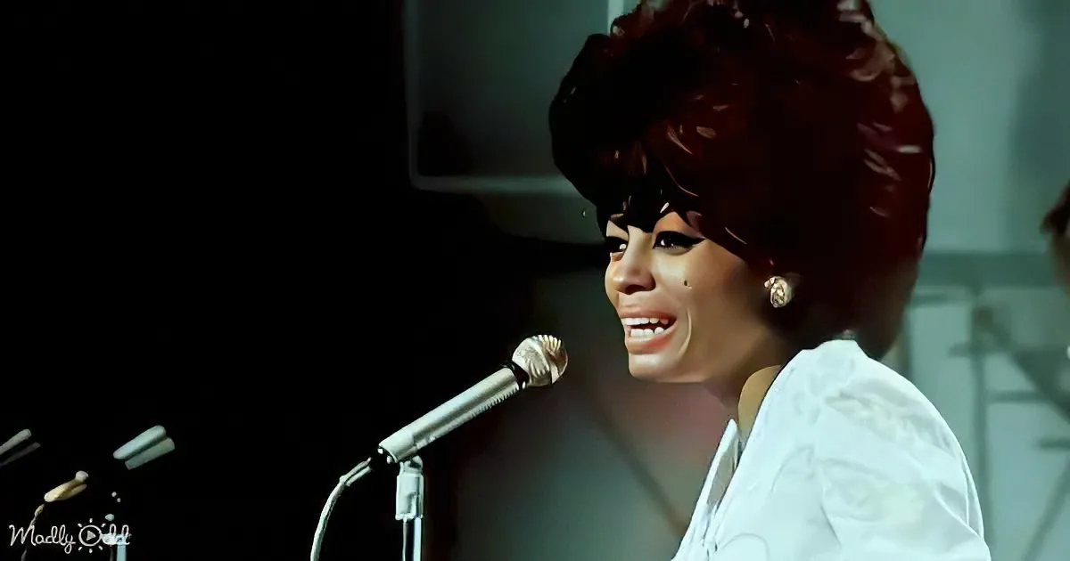 A woman with a large afro hairstyle sings into a microphone, wearing a white outfit.