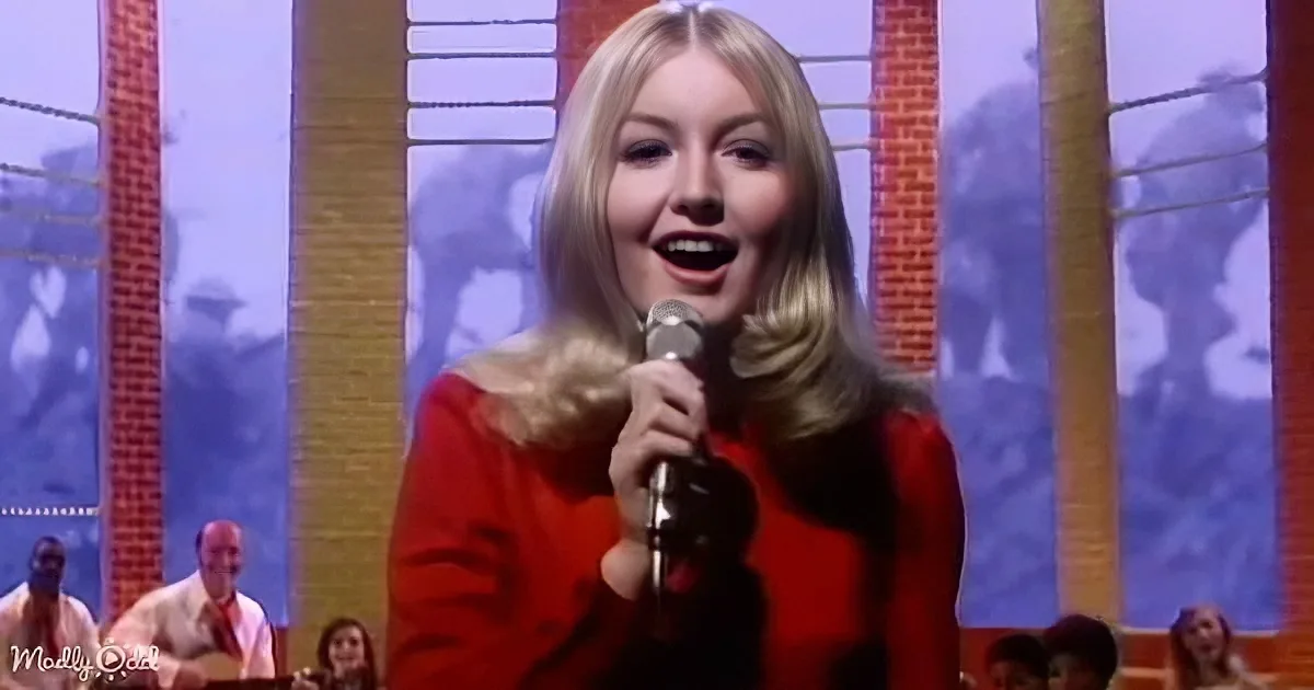 A woman in a red dress sings into a microphone on stage with a brick backdrop and audience.