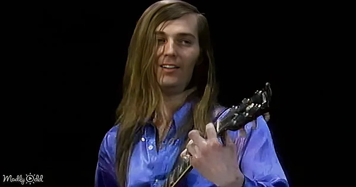 A man with long hair in a blue shirt holds a guitar, appearing to play or sing.