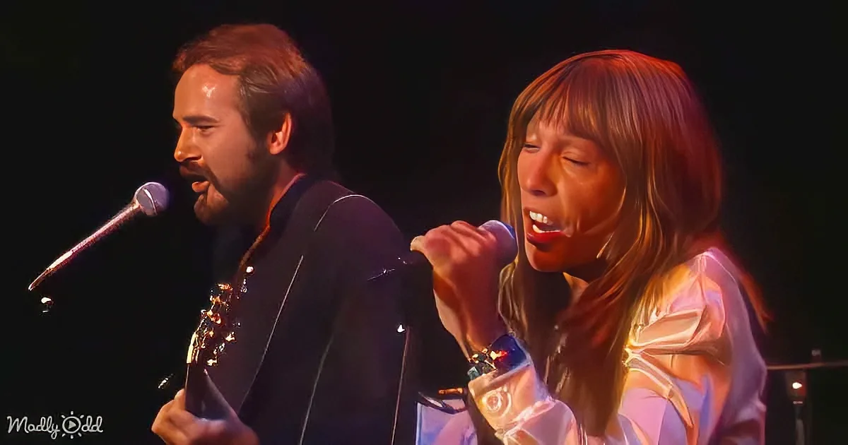 A man and woman sing passionately on stage, the man playing guitar, both illuminated by stage lights.