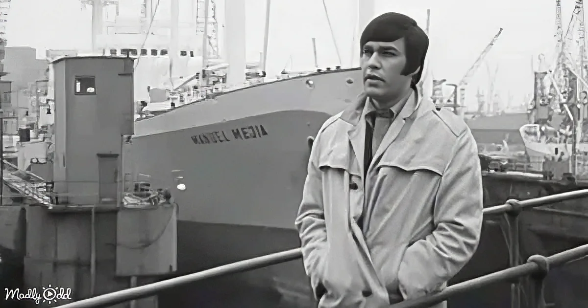 A man in a trench coat stands on a dock, looking out over a shipyard with a large ship labeled "Manuel Media" in the...