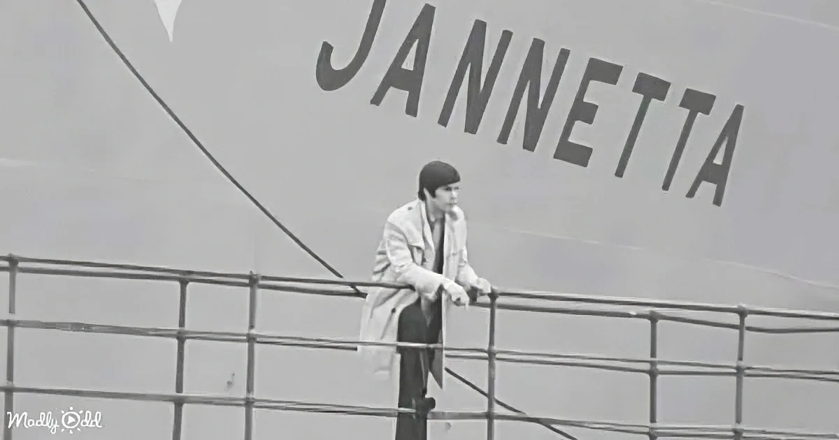 A person leans on a railing, gazing at the ship named "Jannetta" in the background.