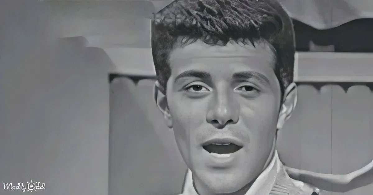 A man with short hair and a white shirt is singing, mouth open, in a black-and-white image.