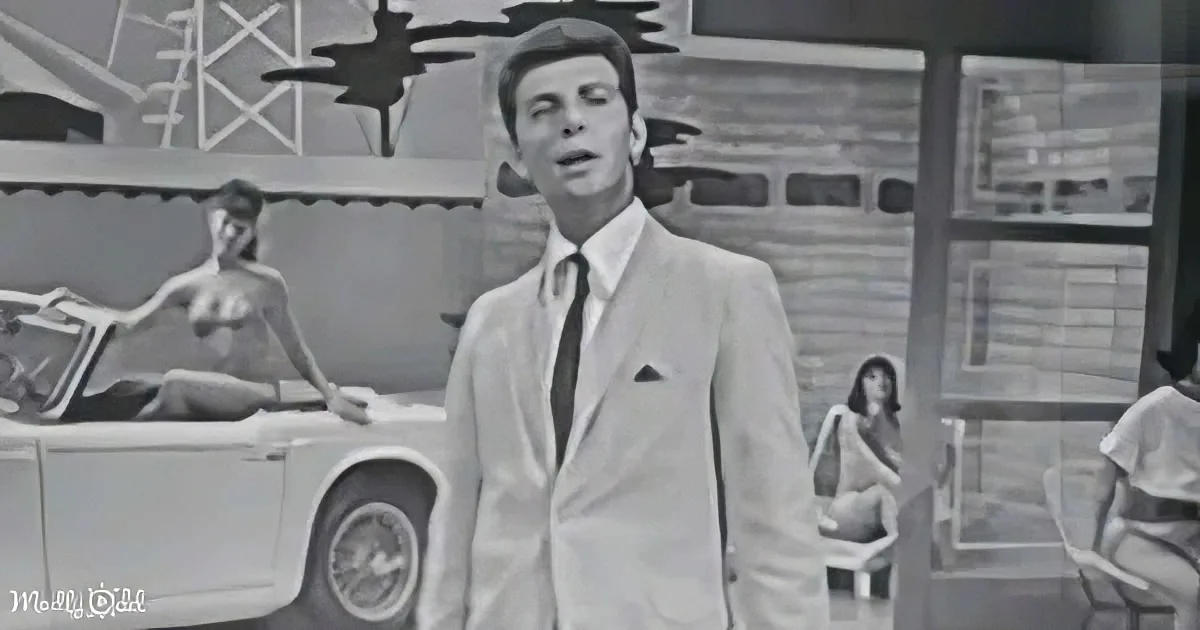 A man in a suit and hat stands in front of a car, with a woman in a bikini and others in the background.