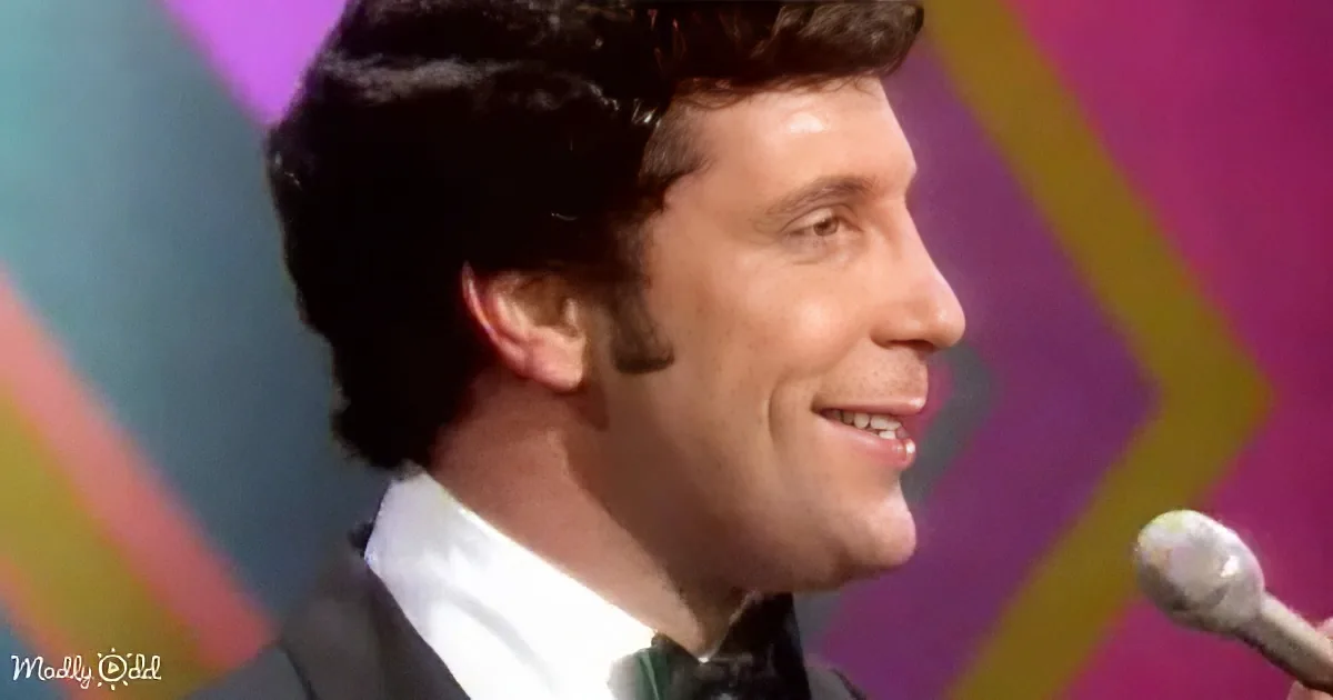 A man in a suit and bow tie smiles while holding a microphone, with a colorful background.