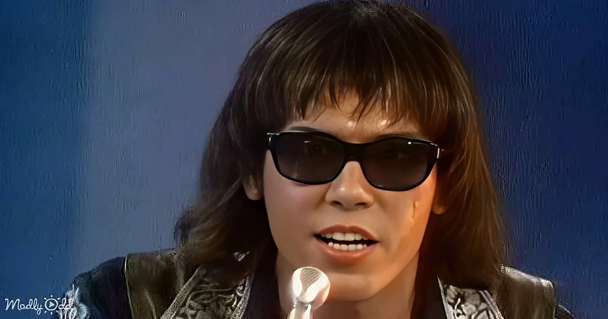 A person with long hair and sunglasses is speaking into a microphone against a blue background.