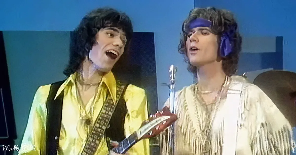 Two individuals in vibrant 70s attire perform energetically, one playing guitar, the other singing into a microphone.
