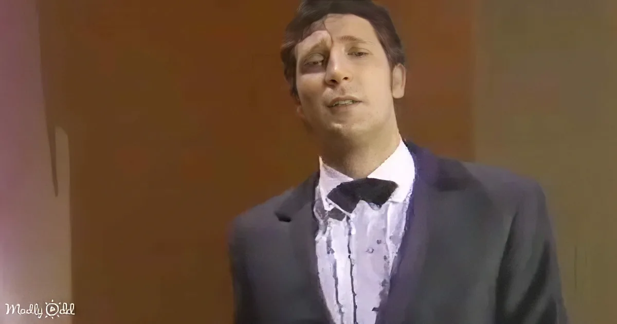 A man in a suit and bow tie stands against a brown background, looking slightly upward.