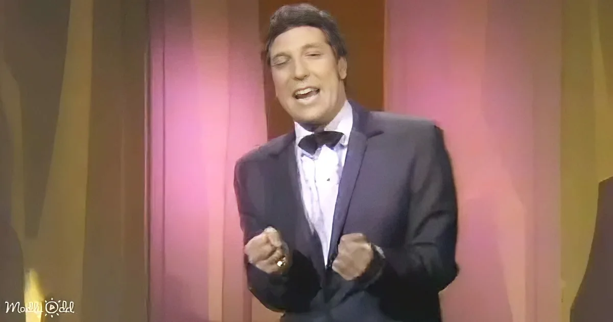 A man in a tuxedo with a bow tie stands with clenched fists, appearing to sing or speak passionately.