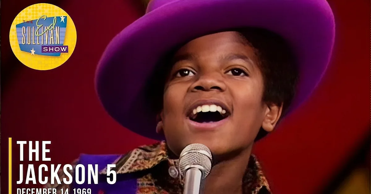 A young person wearing a vibrant purple hat and colorful outfit smiles into a microphone against a red backdrop.