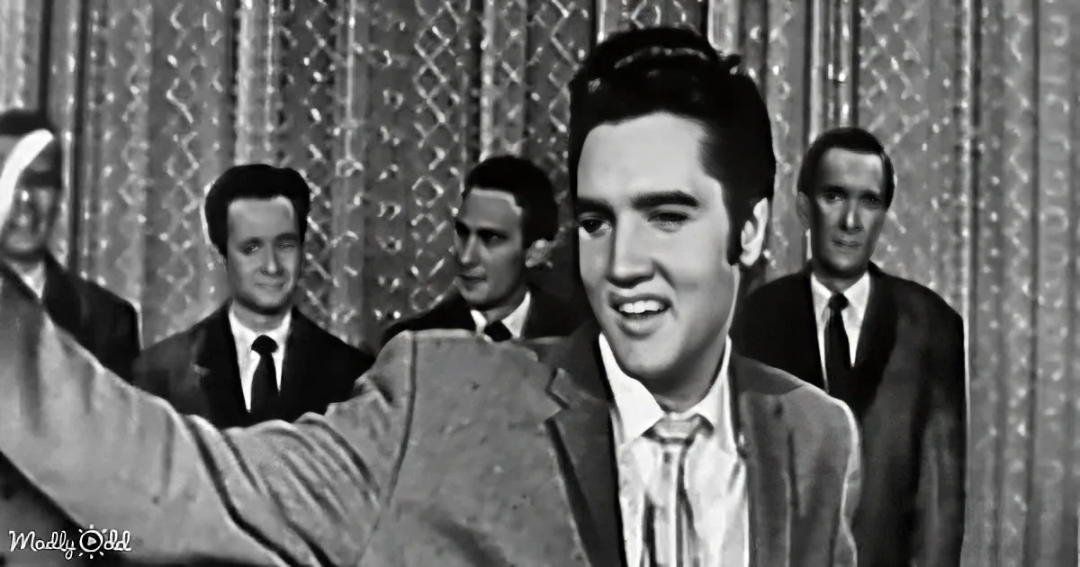 Elvis Presley smiles, raising his arm, with four men in suits behind him.