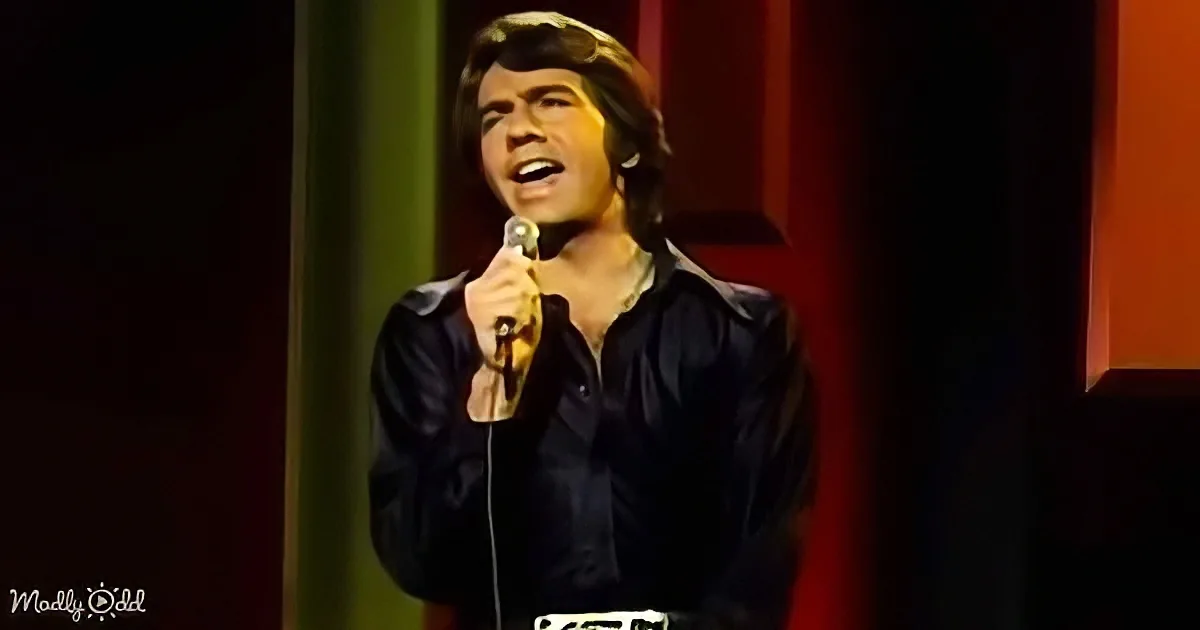 A man in a black shirt sings into a microphone on stage, with a red curtain backdrop.