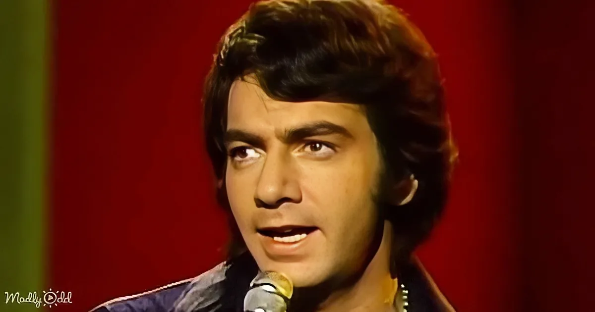 A man with dark hair and a microphone, wearing a dark jacket, appears to be singing or speaking on stage.