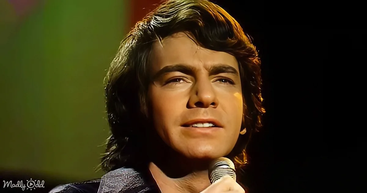 A man with long, dark hair holding a microphone, looking upwards with a serious expression.