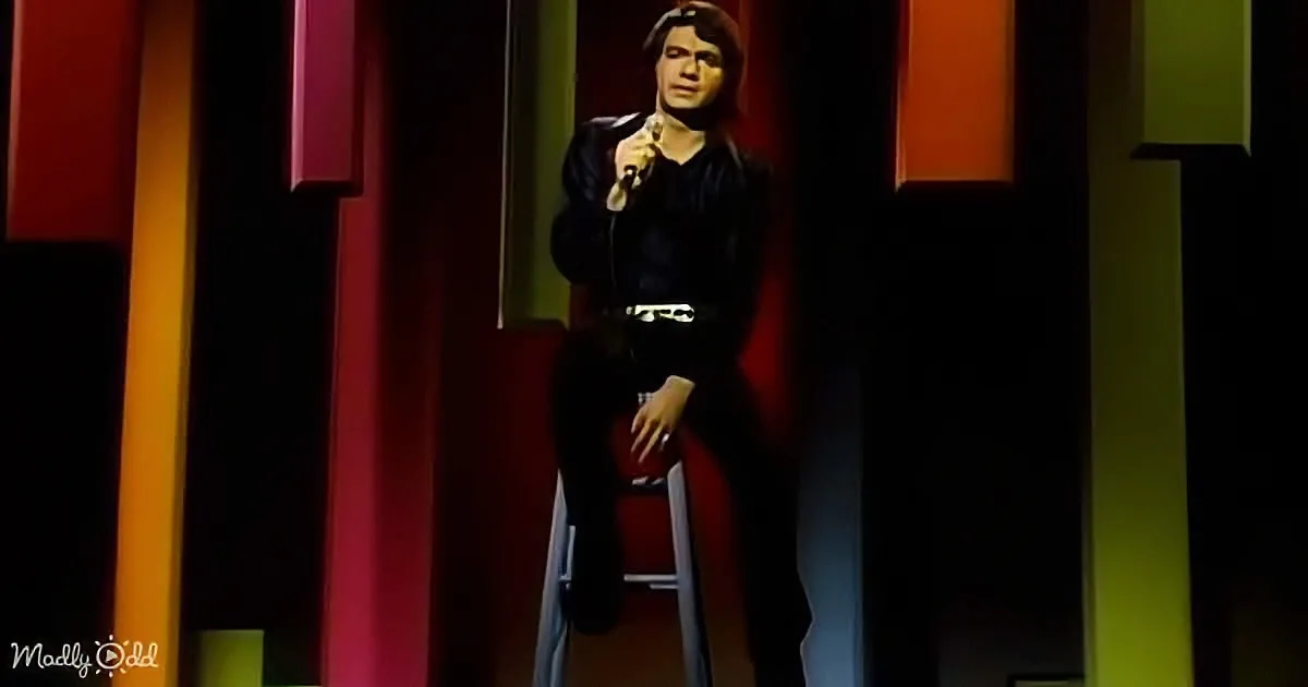 A person in a black outfit sits on a stool against a colorful, geometric background.