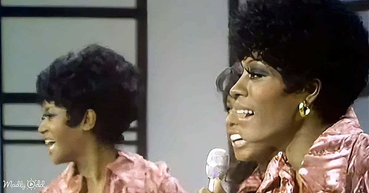A woman with a large afro hairstyle sings into a microphone, wearing a shiny pink outfit.