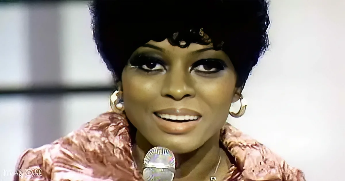 A woman with a short, styled afro hairstyle, wearing a pink textured jacket and large hoop earrings, speaks into a...