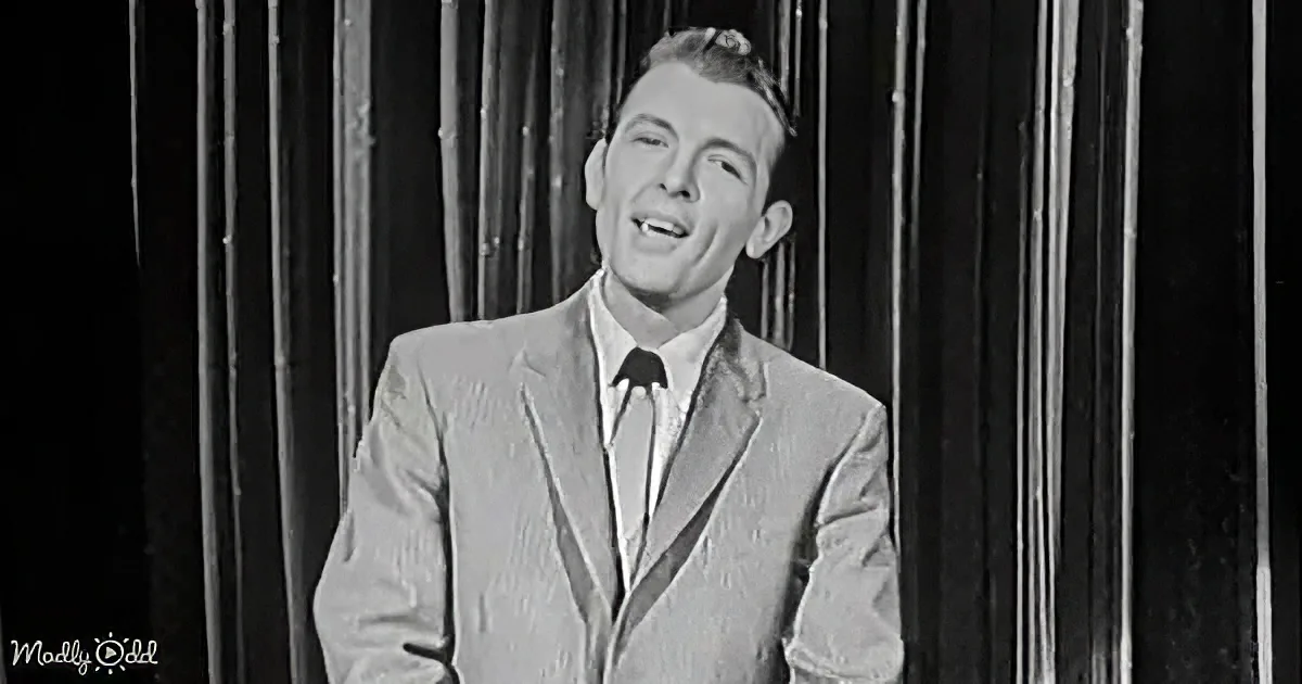 A man in a suit and tie smiles on stage with a curtain backdrop.