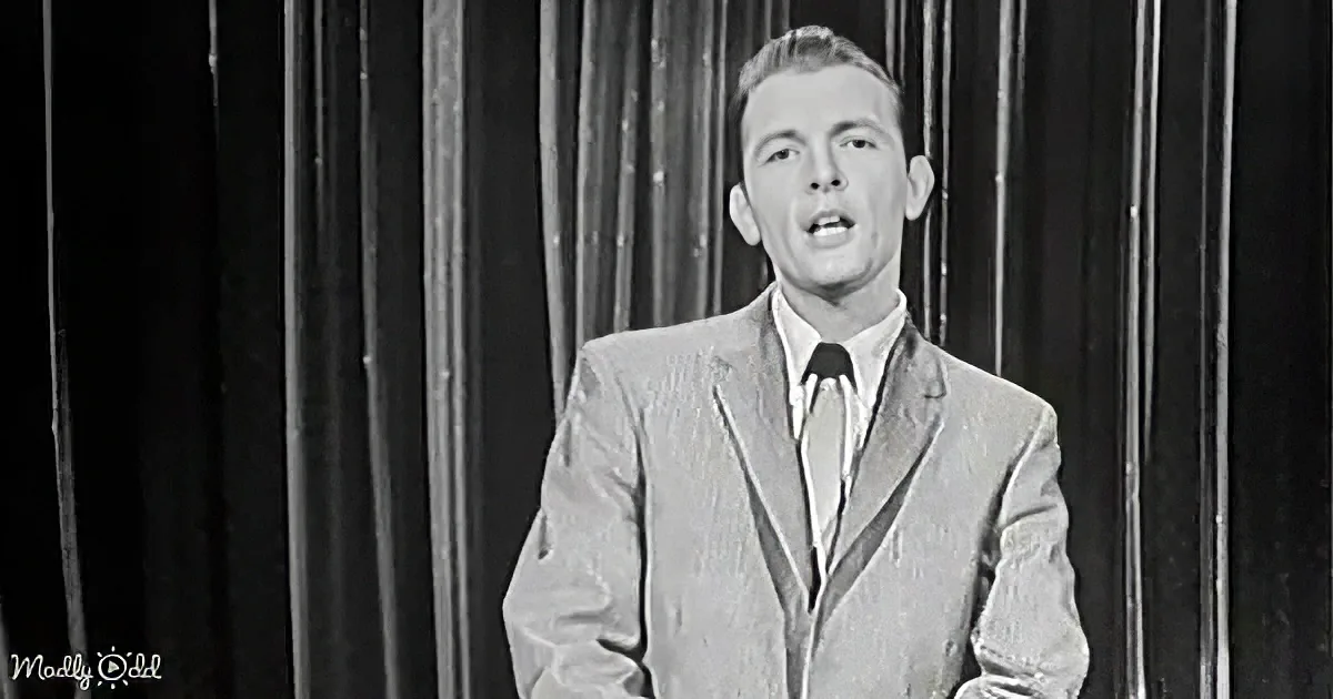 A man in a suit and tie stands on stage, speaking, with a dark curtain backdrop.