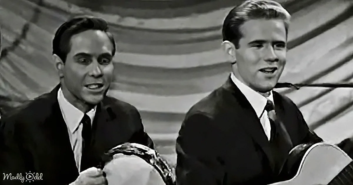 Two men in suits, one playing a guitar, both smiling and singing, with a blurred background.