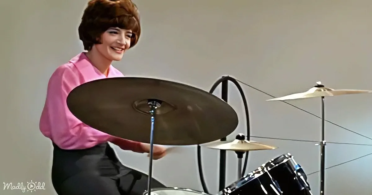 A person in a pink shirt smiles while playing a drum set with a cymbal in hand.