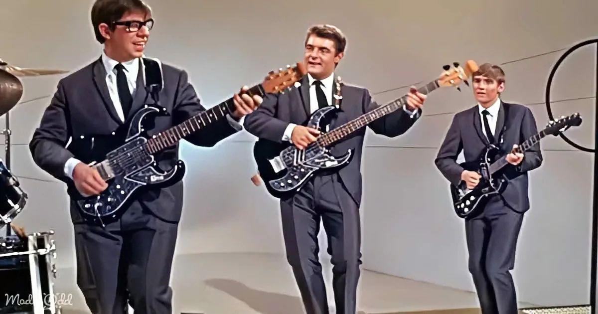 Three men in suits play guitars on stage, one in the center holding a bass.