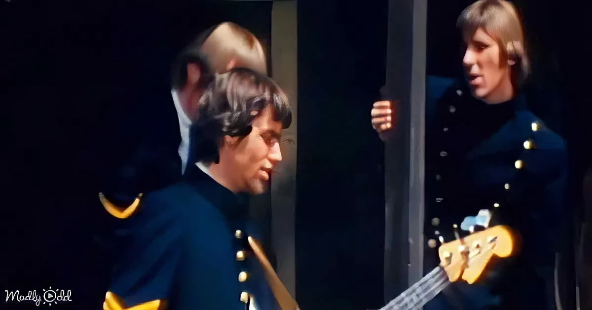 A man in a military-style uniform plays a guitar, with another person in a similar uniform holding a bass guitar in the...