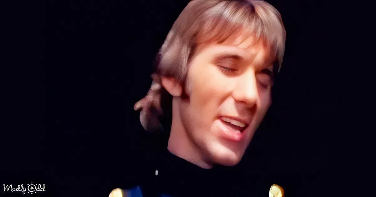 A person with long, light-colored hair and a dark outfit is shown against a dark background.