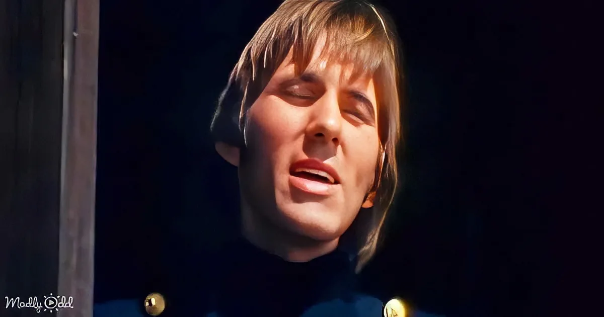 A person with shoulder-length hair, wearing a dark outfit, appears to be singing or speaking with their eyes closed.