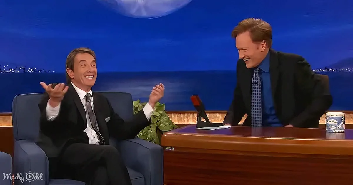 Two men in suits, one seated in a chair, the other behind a desk, both smiling and laughing on a talk show set.