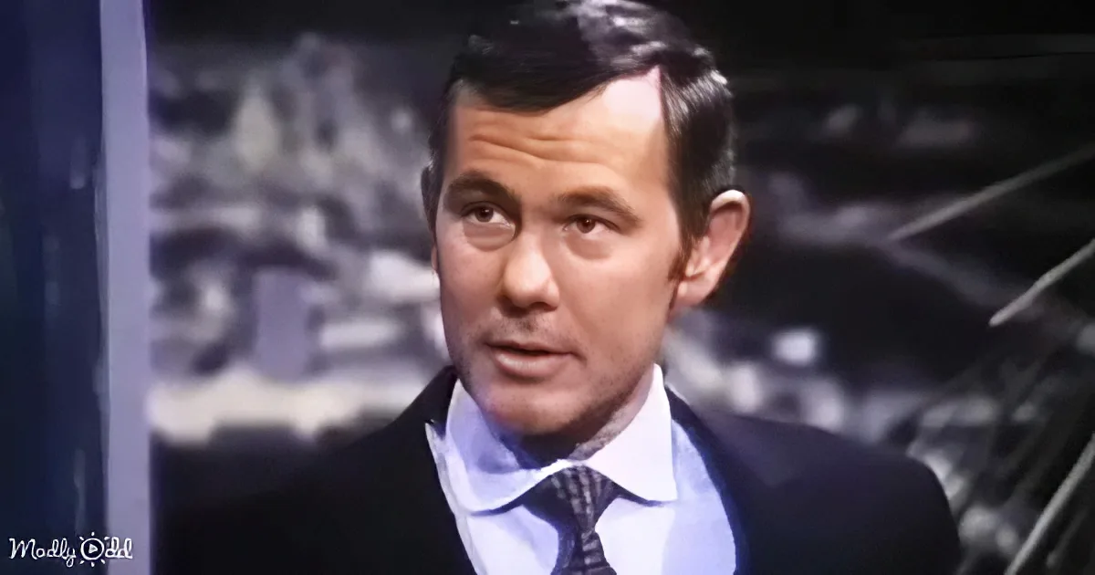 A man in a suit and tie stands in a dimly lit room, looking slightly to the side.
