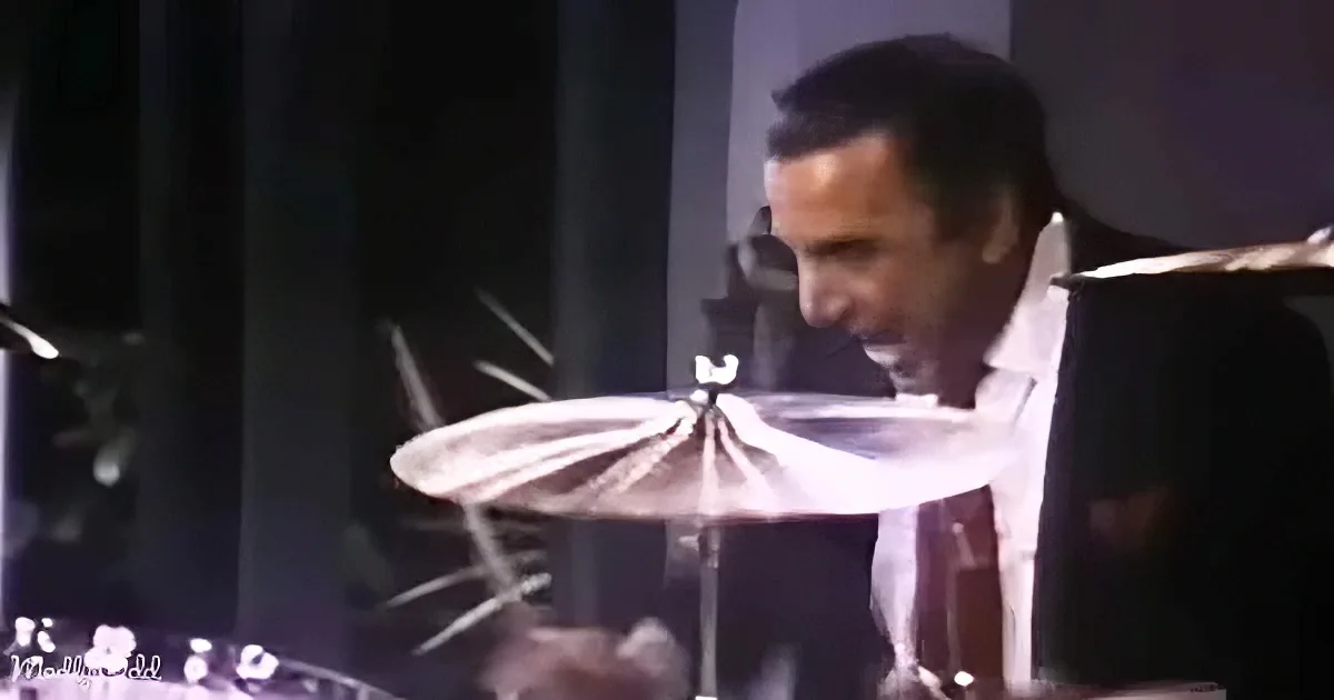 A man in a white shirt and dark suit is playing drums, focusing intently on the cymbal.