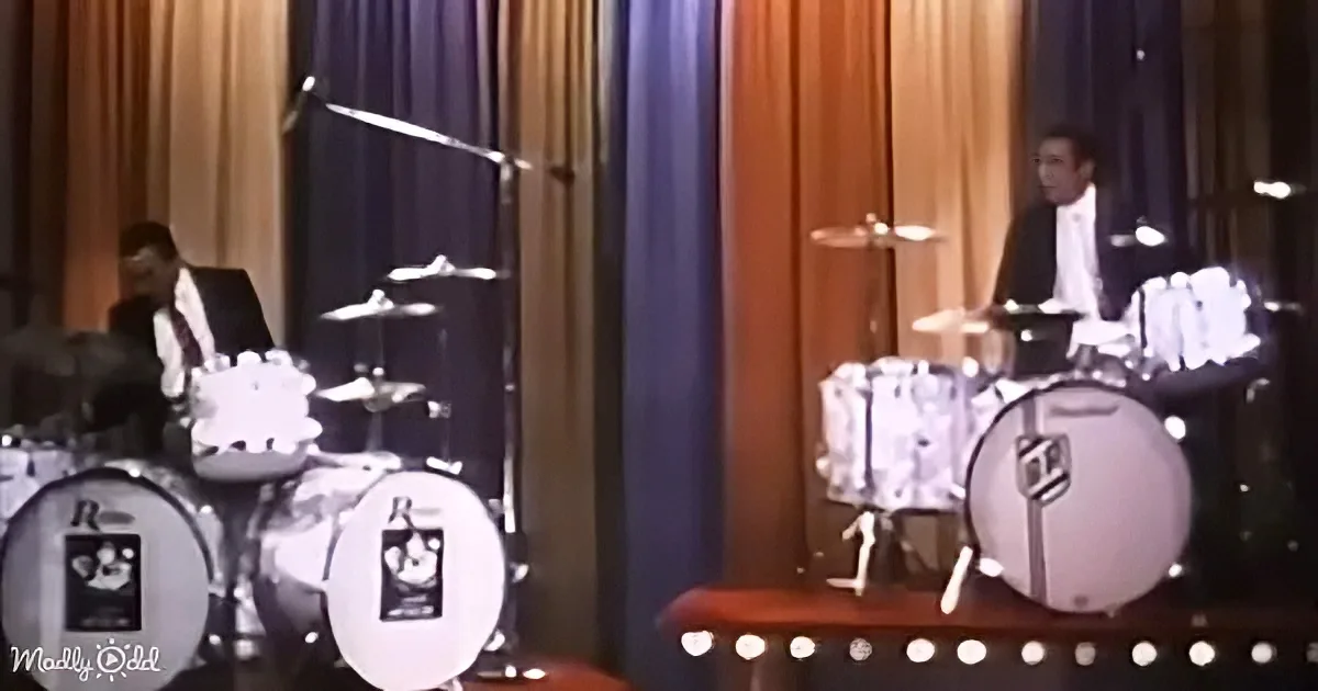 Two drummers in suits play white drums on a stage with wooden paneling and curtains.