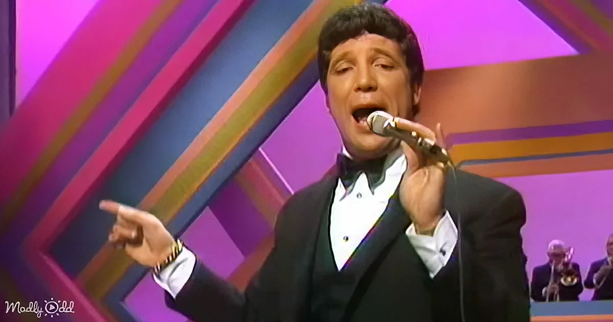 A man in a tuxedo sings into a microphone, pointing to the left, with a colorful geometric background.