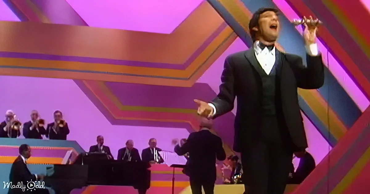 A man in a tuxedo sings passionately on stage with a colorful geometric backdrop and musicians in the background.