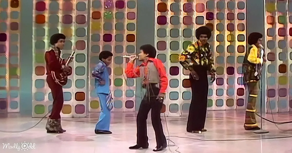 A group of musicians in colorful outfits perform on stage with a vibrant geometric backdrop.