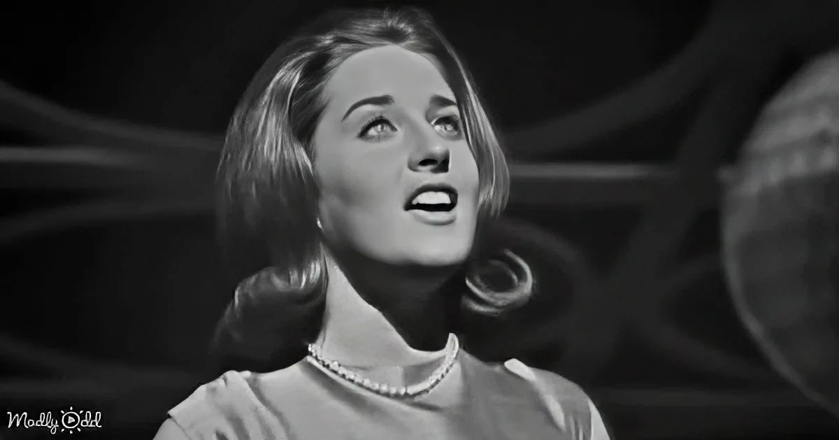 A woman with short hair and a pearl necklace looks upwards, possibly singing, in a black-and-white image.