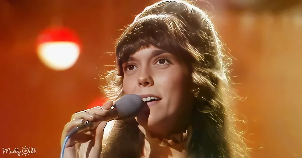 A person with long, wavy hair sings into a microphone against a warm, orange background.