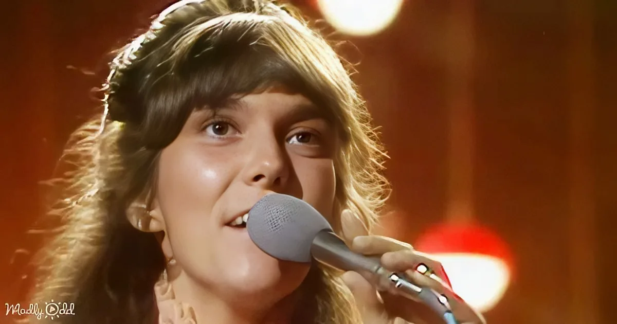 A person with long, wavy hair sings into a microphone, illuminated by stage lights.