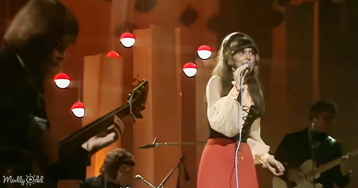 A woman sings into a microphone on stage, surrounded by musicians and red-and-white lights.