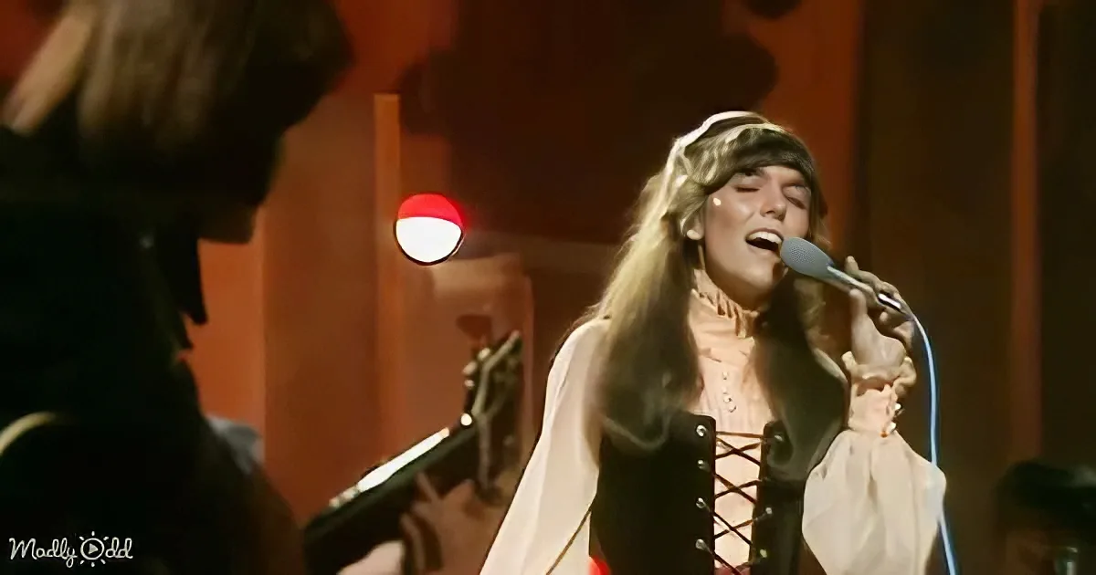A woman in a white and black outfit sings into a microphone on stage, with a guitarist in the background.