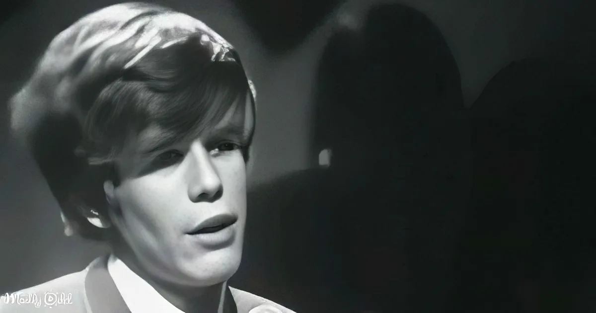 A person with short, styled hair and a white shirt is shown in a close-up, with a dark background.