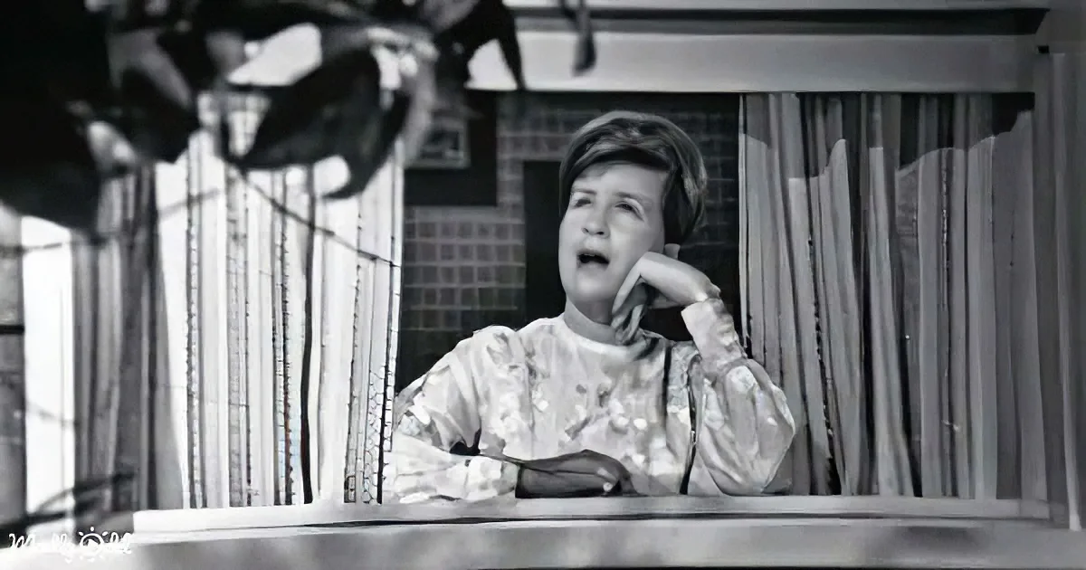 A person in a patterned outfit sits at a desk, hand on chin, in a room with curtains and a window.