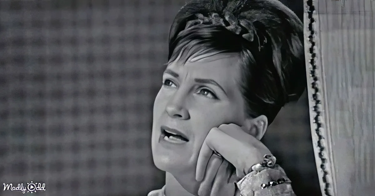 A woman with short hair, wearing a headband, looks up with a hand on her chin, in a black-and-white image.