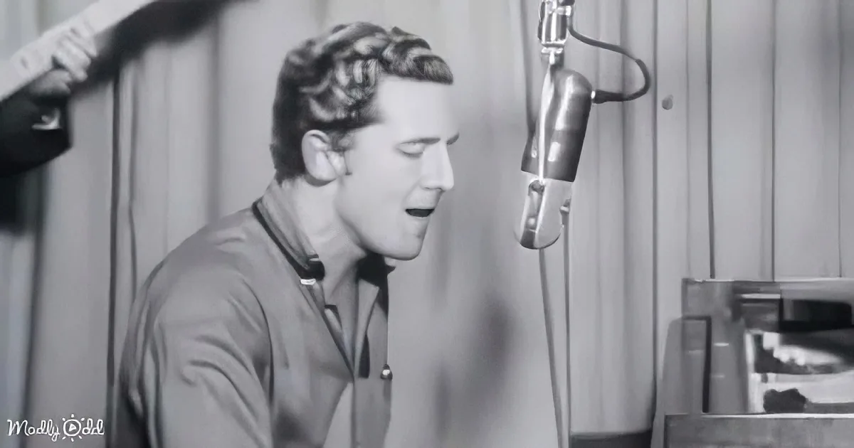 A man sings into a microphone in a recording studio, wearing headphones and a casual shirt.