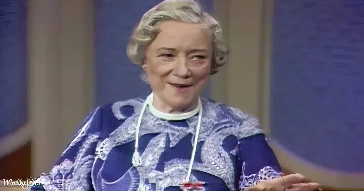 An older woman with gray hair, wearing a blue and white patterned outfit, smiles while holding a small object in her hands.