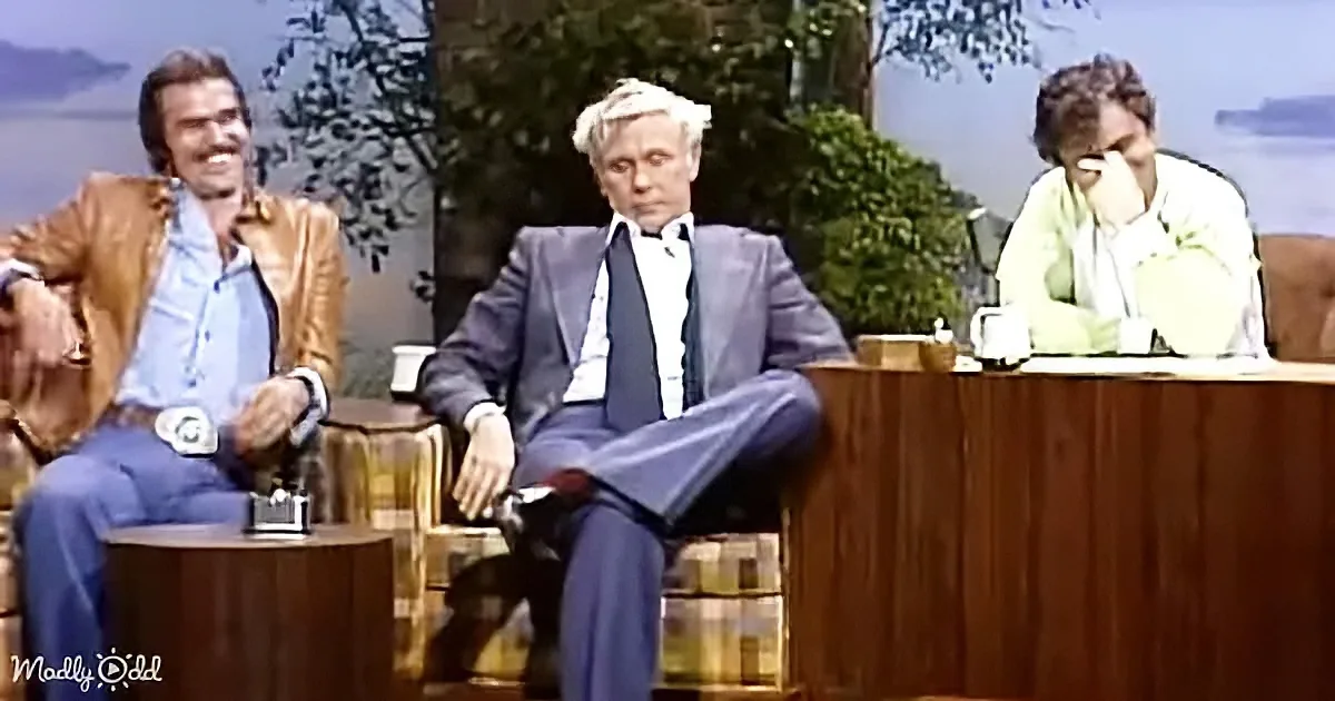 A man in a gold jacket sits on a couch, smiling, with another man in a suit and a woman in a green shirt behind him.