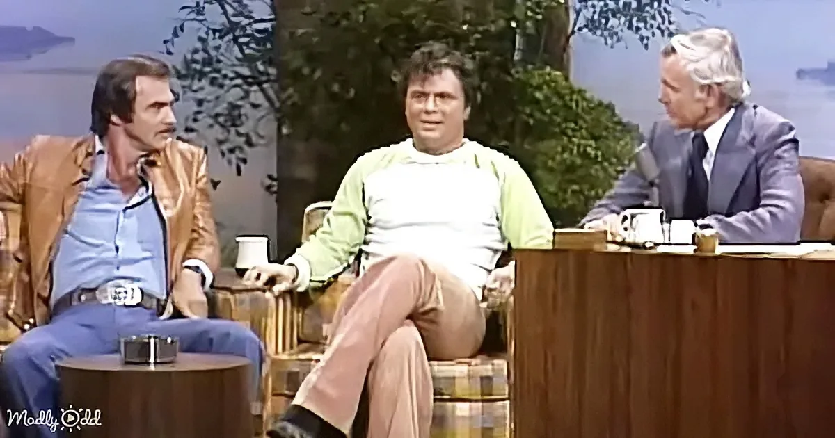A man in a green shirt sits between two others on a talk show set with a scenic backdrop.